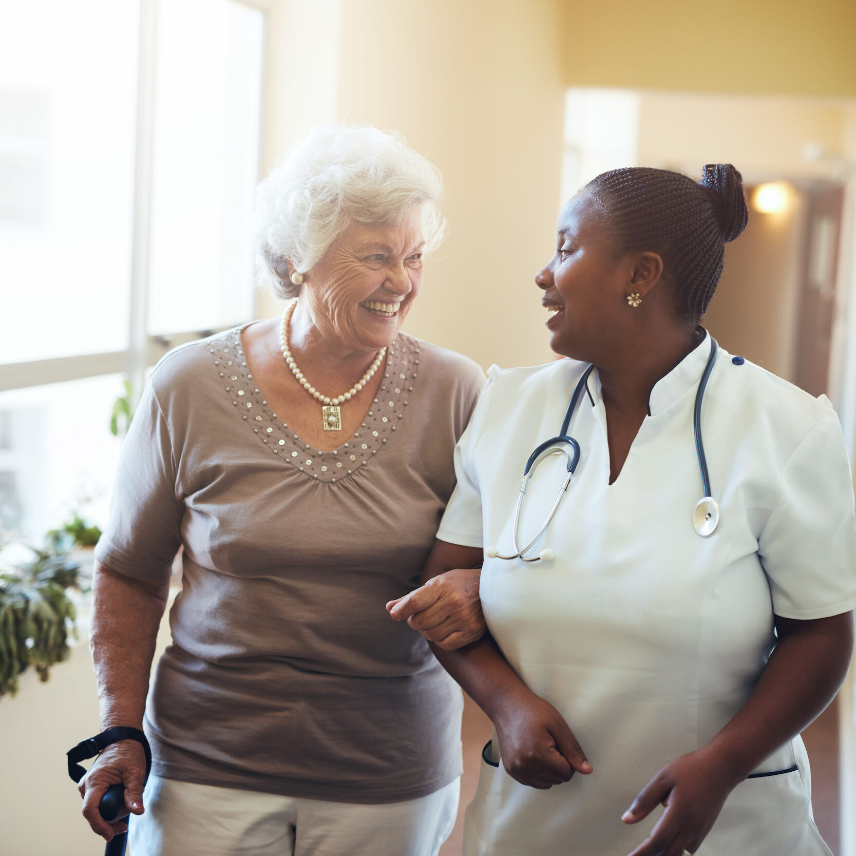 A senior woman and a nurse walk arm-in-arm down a hallway, smiling and talking. The nurse wears a stethoscope and white uniform, while the woman uses a cane. Sunlight streams through the windows beside them. A senior woman and a nurse walk arm-in-arm down a hallway, smiling and talking. The nurse wears a stethoscope and white uniform, while the woman uses a cane. Sunlight streams through the windows beside them.